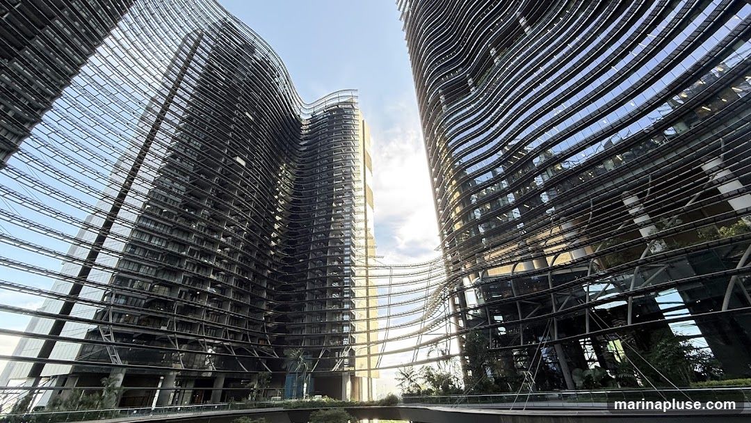 A low-angle shot captures the striking architecture of two modern skyscrapers with undulating, wavy facades. The buildings are clad in glass and metal, reflecting the bright sky and sunlight. Greenery is visible on balconies and at the base of the structures, suggesting a blend of urban design with nature. The perspective emphasizes the scale and unique design of the buildings.