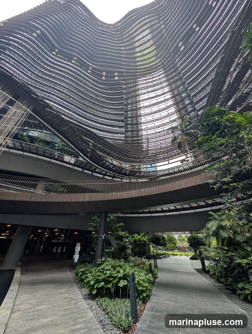A low-angle shot looking up at a uniquely designed modern building with curved, layered facades. Lush green plants and trees are integrated into the architecture, growing on balconies and along walkways. The pathways are paved with stone, and the overall scene suggests a blend of urban design and nature.