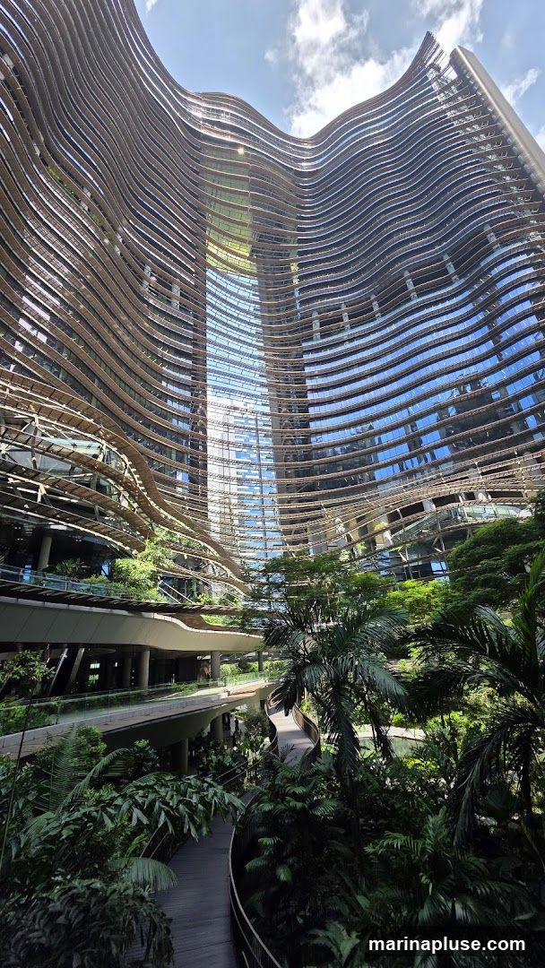 A striking modern building with a wavy facade made of horizontal wooden slats and reflective glass. The interior features multiple levels with walkways winding through dense tropical foliage, including palm trees and various plants. The architecture creates a sense of an urban oasis.