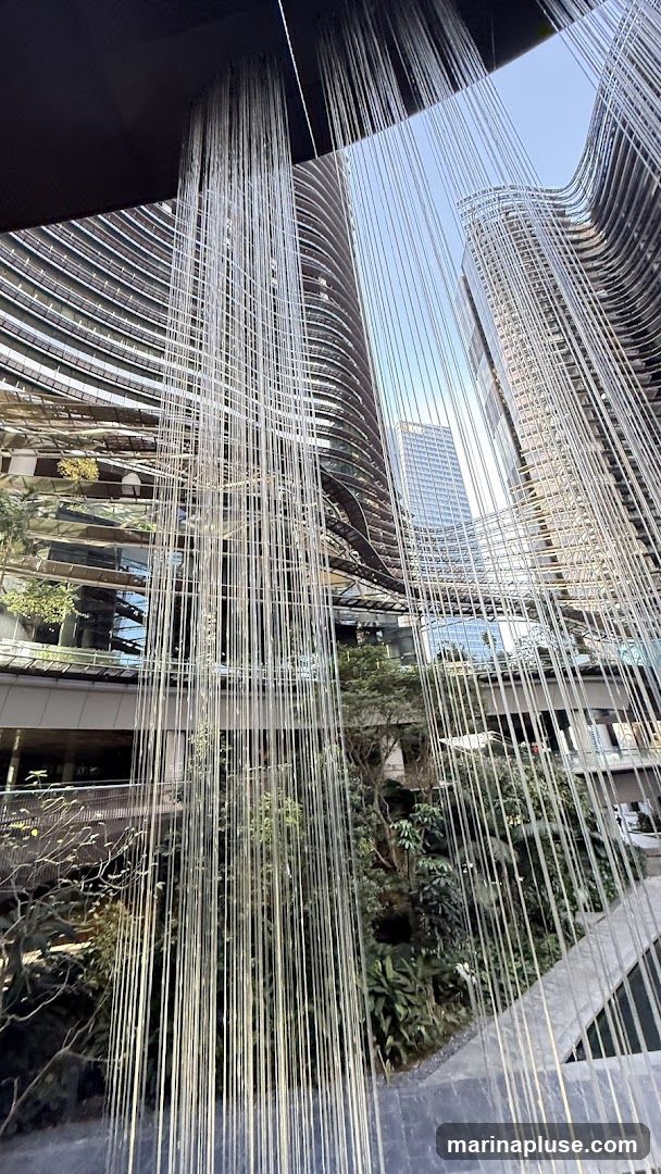 A view looking up at modern, curved buildings with many windows, partially obscured by numerous thin, vertical, light-colored strands hanging from above. Lush greenery is visible on lower levels and balconies. The sky is clear and blue.