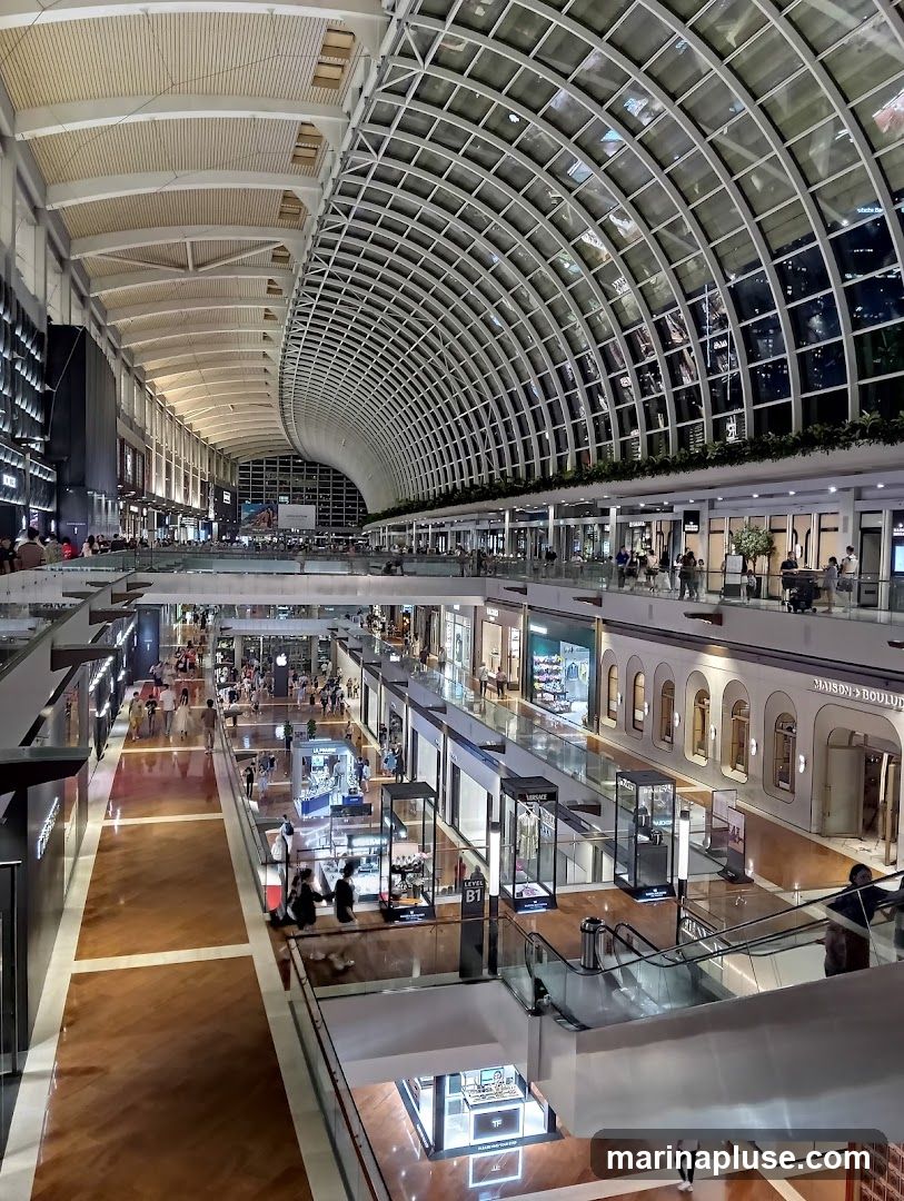 A wide-angle view of the interior of a large, modern shopping mall. The architecture features a distinctive curved, grid-like ceiling with glass panels, allowing natural light to filter in. Below, multiple levels of shops and walkways are visible, bustling with shoppers. Escalators connect the different floors, and display cases showcase merchandise. The overall impression is one of a spacious and upscale retail environment.