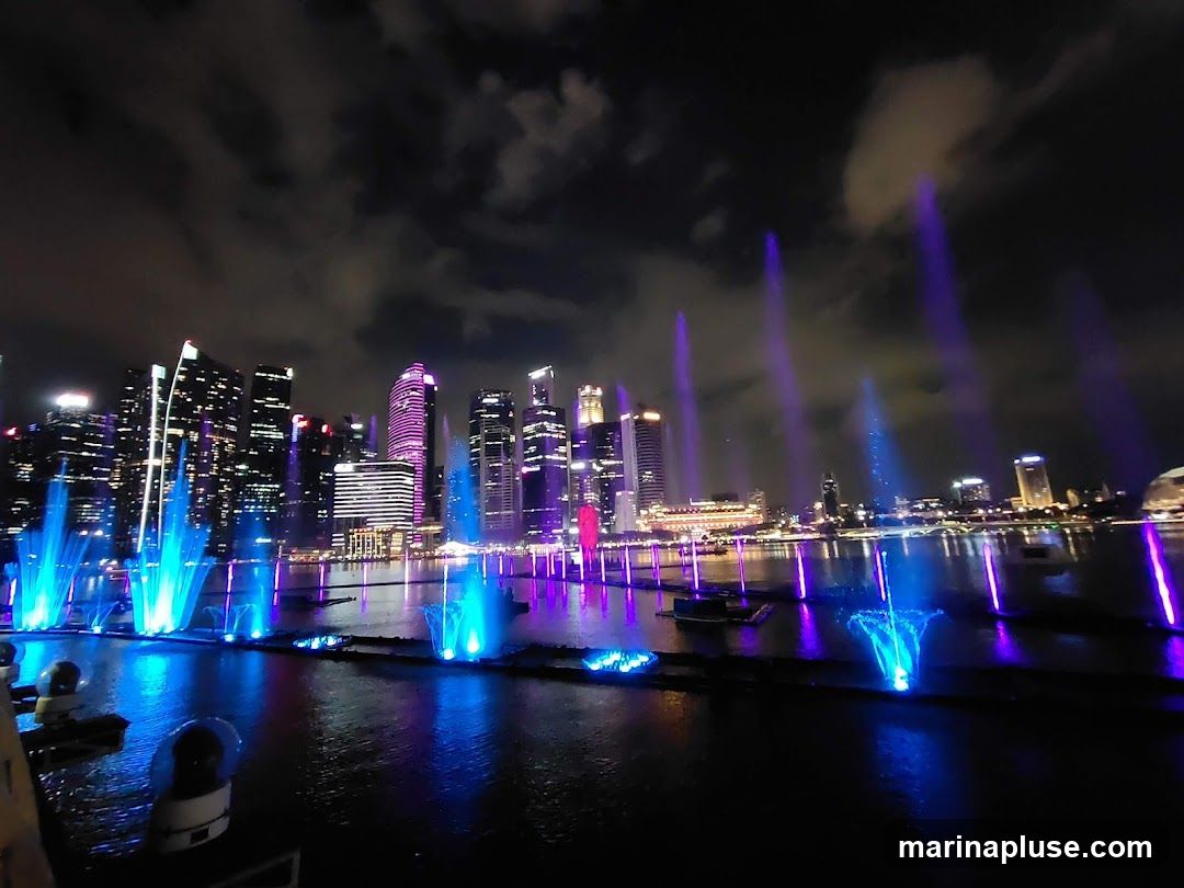 A vibrant nighttime scene featuring a water fountain show with colorful lights, set against a backdrop of a modern city skyline. The fountains spray water high into the air, illuminated by blue and purple lights, creating a dazzling spectacle on the water's surface. The city buildings in the background are lit up, reflecting on the water. The sky is dark with scattered clouds.