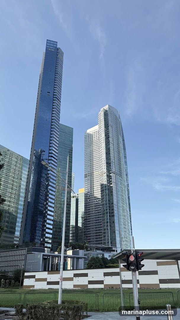 A low-angle view of several modern skyscrapers against a clear blue sky with scattered clouds. The buildings are characterized by their glass facades, reflecting the sky and surrounding environment. In the foreground, a traffic light shows a red signal, and a bus stop shelter is visible. A low wall with a checkered pattern of brown and beige panels runs along the middle ground, with a strip of green grass and bushes in front of it.