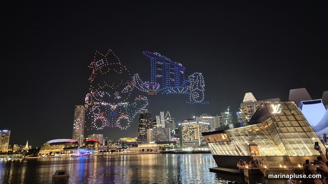 A nighttime cityscape with a drone light show forming various shapes in the sky. In the foreground, a modern glass building with a prominent Louis Vuitton logo is visible. The city skyline and a body of water with reflections of lights are also present.