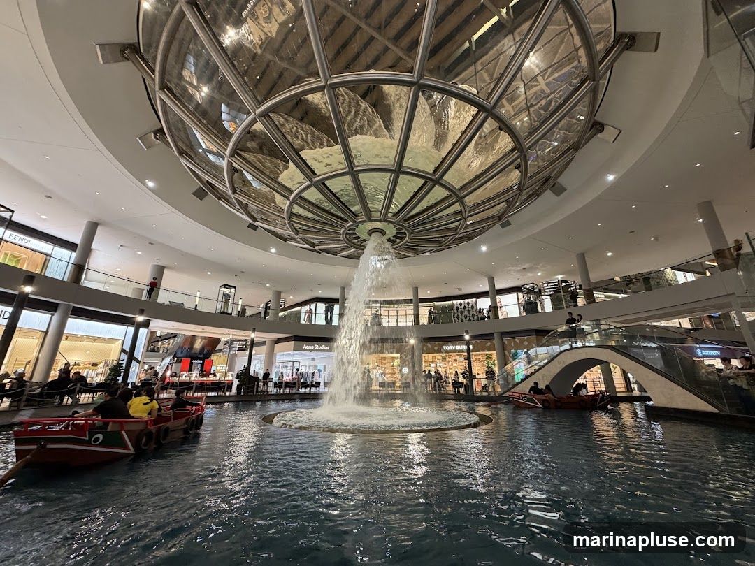 An indoor shopping mall features a large, circular water feature in the center, with boats carrying passengers. Above the water, a large, ornate glass and metal structure is visible, from which a waterfall cascades into the pool. Surrounding the water feature are multiple levels of shops, with escalators and walkways connecting them. The shops have visible signage, including Fendi, Tory Burch, and Acne Studios. People are seen browsing in the shops and riding in the boats.