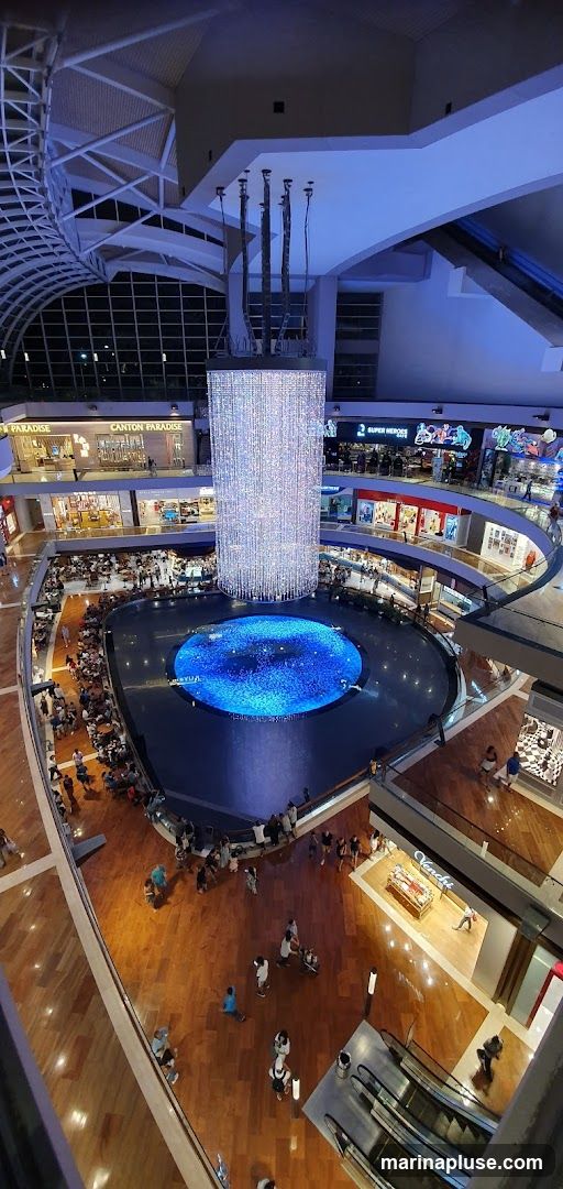 An aerial view of a large, modern shopping mall atrium. In the center is a circular pool of water with a blue light display, surrounded by a wide walkway. Above the pool hangs a massive, cylindrical chandelier made of numerous sparkling lights. Multiple levels of shops and restaurants are visible around the atrium, with shoppers browsing and dining. Escalators and walkways connect the different levels. The architecture features a high, curved ceiling with exposed beams and a large glass facade.