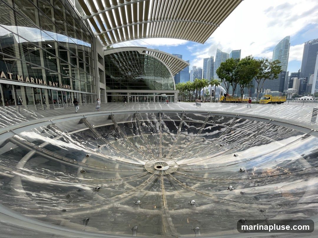 A wide shot of the exterior of Marina Bay Sands, with a large, clear, circular structure in the foreground. The building's glass facade reflects the sky and surrounding cityscape. People are visible walking in front of the building. In the background, a line of modern skyscrapers and palm trees are seen under a cloudy sky. Two yellow trucks are parked on the right side of the image.