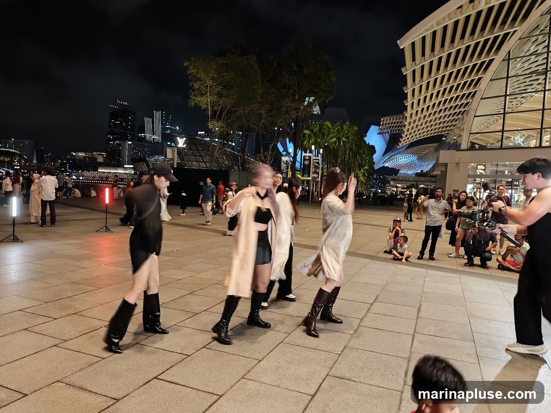 A group of people are filming a dance performance on a paved plaza at night. Several women in fashionable outfits are dancing, while a man operates a video camera. Other people are watching the performance or walking by. In the background, there are modern buildings with distinctive architecture and city lights. A large logo, possibly for Louis Vuitton, is visible on one of the buildings.