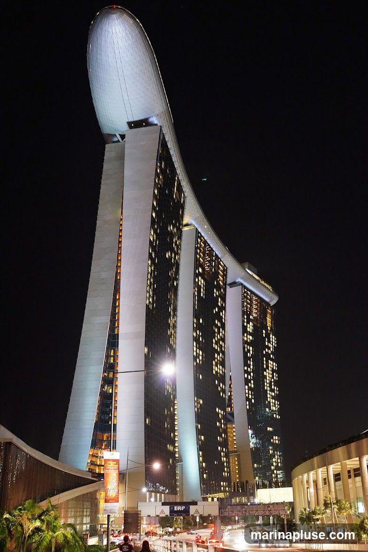 A nighttime view of the iconic Marina Bay Sands hotel in Singapore, showcasing its distinctive architecture with three towers topped by a large, boat-shaped structure. The building is illuminated, with many windows lit up, suggesting occupancy. The surrounding area shows some streetlights, traffic, and greenery. A banner is visible on the lower left, but its text is not fully legible.