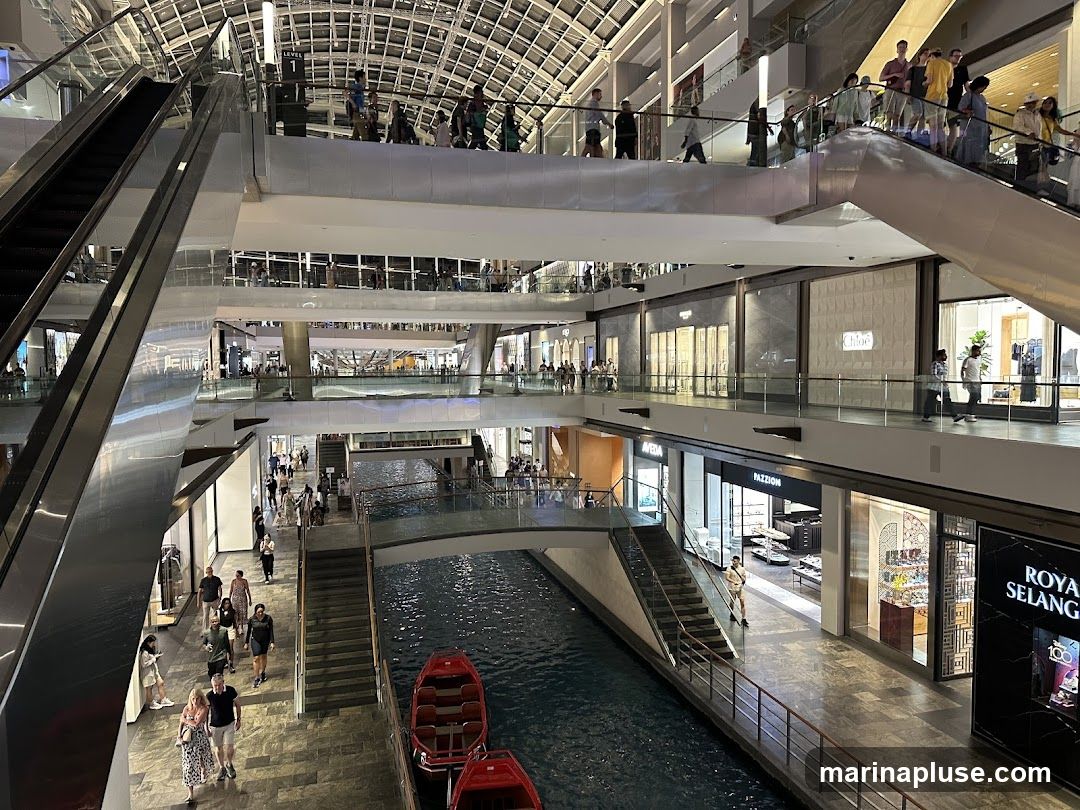 The image shows the interior of a multi-level shopping mall with escalators, shops, and a canal running through the center. People are seen walking on the different levels and on a bridge over the canal. Red boats are docked in the canal. Shops visible include Chloe, Aveda, Pazzion, and Royal Selangor. The architecture is modern with a large, arched glass ceiling.