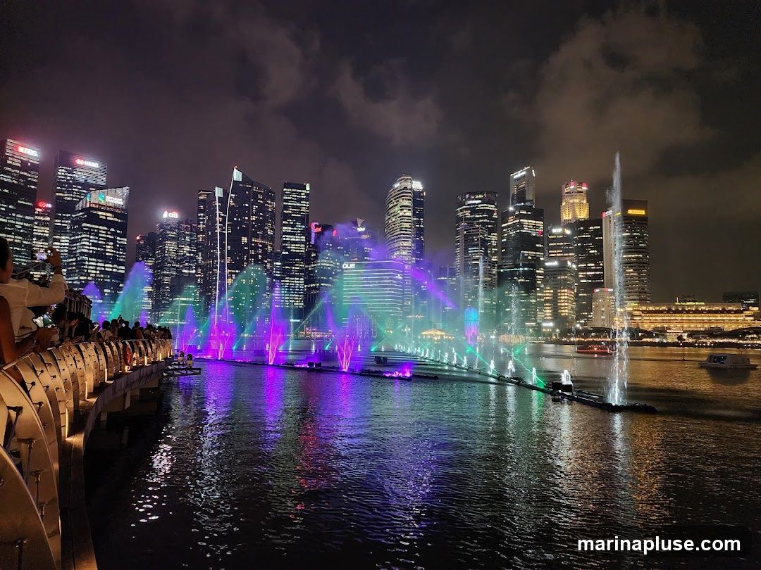 A vibrant nighttime scene featuring a water fountain show with colorful lights (purple and green) set against a backdrop of a modern city skyline. People are visible on a walkway in the foreground, observing the spectacle. The water reflects the lights and the surrounding buildings.