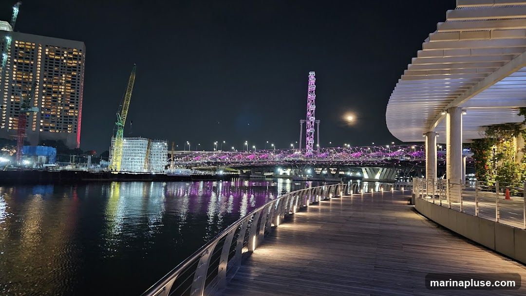 A wooden promenade curves along a dark river at night. The promenade is illuminated by lights along its railing and under its canopy. Across the river, a brightly lit bridge with purple accents spans the water, and a Ferris wheel-like structure glows pink. In the background, a tall building with many illuminated windows stands next to a construction site with cranes. The moon is visible in the dark sky.