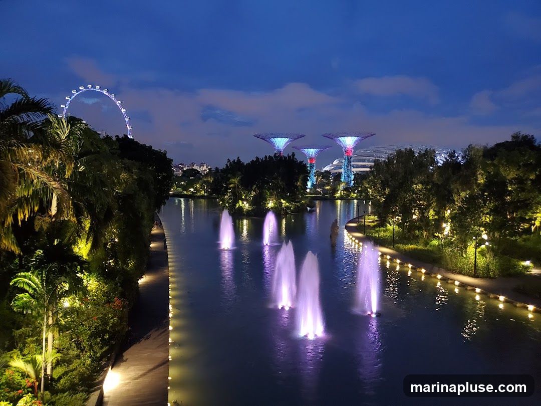 A scenic nighttime view of Gardens by the Bay in Singapore. The image features illuminated Supertrees, a Ferris wheel in the distance, and fountains spraying water in a canal, all set against a twilight sky. Lush greenery and pathways are lit by warm lights, creating a magical atmosphere.