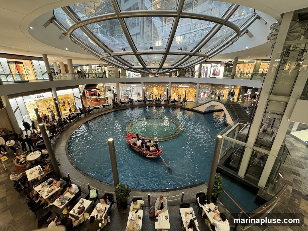 An aerial view of a large indoor shopping mall featuring a central canal with a boat carrying passengers. Shops with illuminated storefronts, including Fendi and Gucci, are visible on multiple levels. People are seated at tables outside cafes and restaurants surrounding the canal. The mall has a modern architectural design with a glass ceiling and escalators.