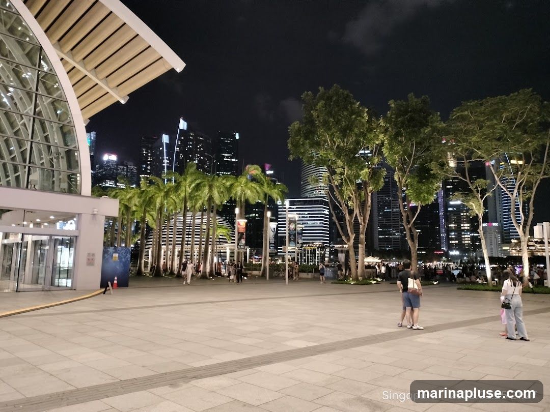 A wide shot of a plaza at night with a modern building on the left and a cityscape with illuminated skyscrapers in the background. Palm trees and other trees are scattered throughout the plaza. Several people are walking and standing around. The image is dated February 25, 2023.
