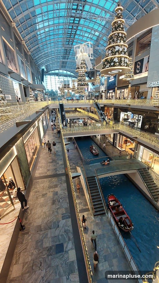 An interior view of a multi-level shopping mall decorated for Christmas. Large, tiered Christmas trees adorned with lights and ornaments hang from the ceiling. A canal with gondola-like boats runs through the center of the mall, with shoppers walking along the elevated walkways. Storefronts with visible signage line the sides of the mall. The ceiling is a glass structure, allowing natural light to filter in.
