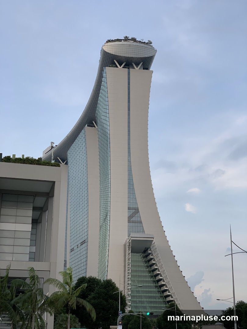 A view of the iconic Marina Bay Sands hotel in Singapore, with its distinctive three towers connected by a large rooftop deck. The building features a modern architectural design with extensive glass facades and a curved, boat-like structure at the top. Lush greenery is visible on the rooftop and surrounding areas. The sky is partly cloudy.