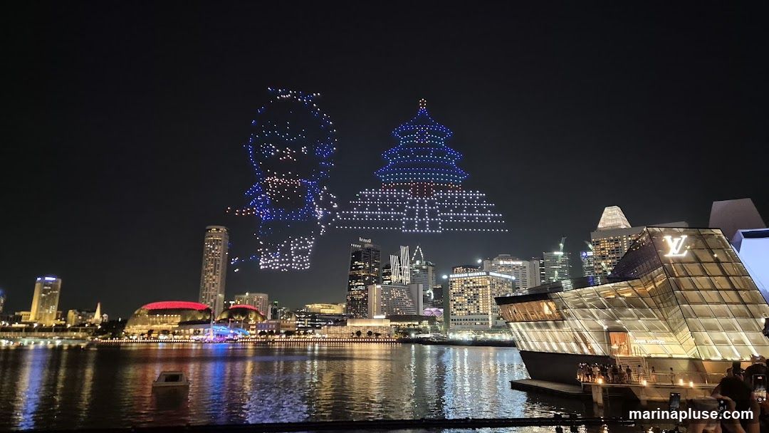 A nighttime cityscape with a drone light show forming images of a character and a pagoda in the sky. In the foreground, a modern glass building with a prominent Louis Vuitton logo is visible. The city skyline features illuminated skyscrapers and a waterfront with reflections in the water.