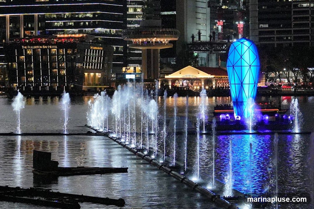 A nighttime view of a waterfront with a large fountain display. In the background, the Fullerton Bay Hotel is visible, along with other illuminated buildings and a distinctive blue, geometric sculpture. The water is lit by the fountains and reflections from the surrounding lights.