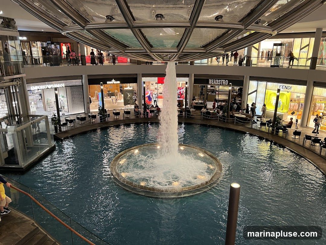 An indoor view of a multi-level shopping mall featuring a large circular fountain in the center. Shops with visible signage such as Emporio Armani, Stone Island, Beanstro, and Fendi are located on the surrounding levels. People are seen browsing in the shops and sitting at tables near the fountain. The architecture includes modern design elements with a glass ceiling structure above the fountain.
