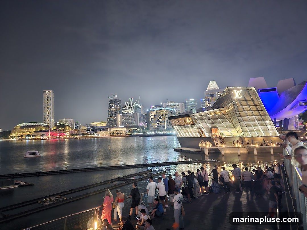 A modern, glass-fronted building with a prominent Louis Vuitton logo on its facade, situated on the waterfront at night. A crowd of people are gathered on a boardwalk in front of the building, with a city skyline visible in the background.