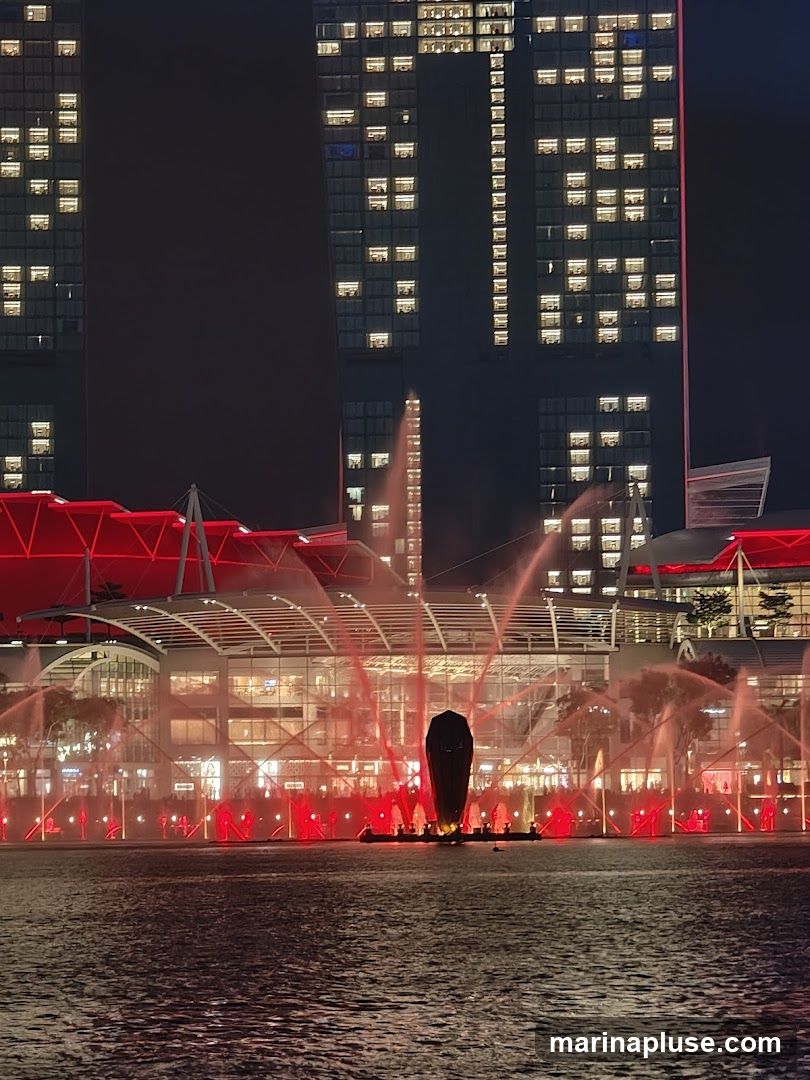 A nighttime scene featuring a water fountain show with red lights and mist, set against a backdrop of illuminated skyscrapers. The fountains are arranged in a line across a body of water, with a large, dark, sculptural element in the center. Behind the fountains, a modern building complex with a red-accented roof structure is visible. The skyscrapers in the background have many windows lit up, creating a grid pattern against the dark sky.