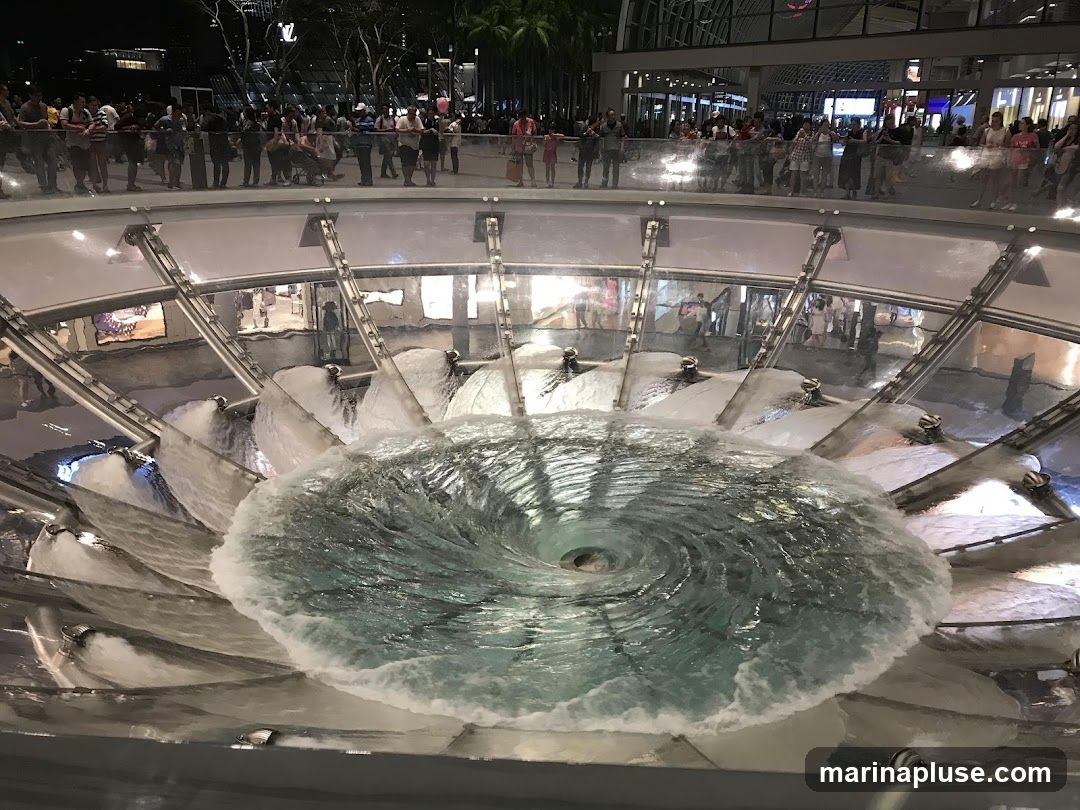 A large, circular fountain with water creating a vortex in the center. Water cascades down from multiple spouts around the perimeter, feeding into the swirling pool. People are visible in the background, standing on a walkway overlooking the fountain. The scene appears to be at night, with artificial lighting illuminating the area.