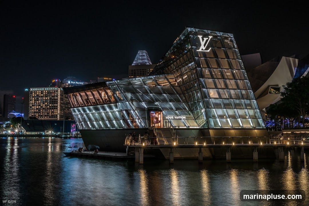 A modern, glass-fronted Louis Vuitton store is illuminated at night, with its distinctive logo prominently displayed on the upper facade. The building, situated on the water's edge, reflects the lights of the surrounding cityscape and the water itself. Stairs lead up to the entrance, where the Louis Vuitton name is visible.