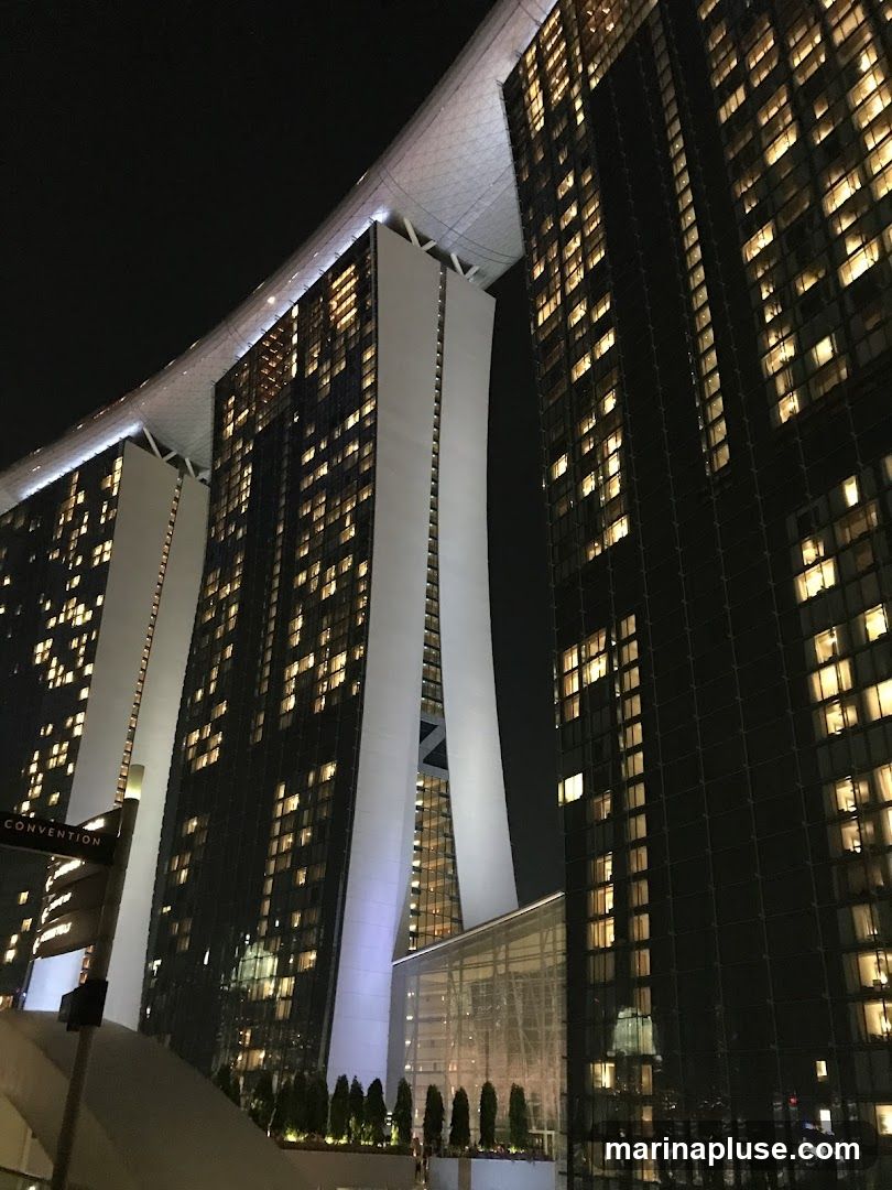 A nighttime view of the iconic Marina Bay Sands hotel in Singapore, showcasing its distinctive architecture with three towers connected by a sky-high platform. The buildings are illuminated with numerous lights from their windows, creating a striking contrast against the dark sky. A sign indicating "CONVENTION" is visible on the left side of the image.