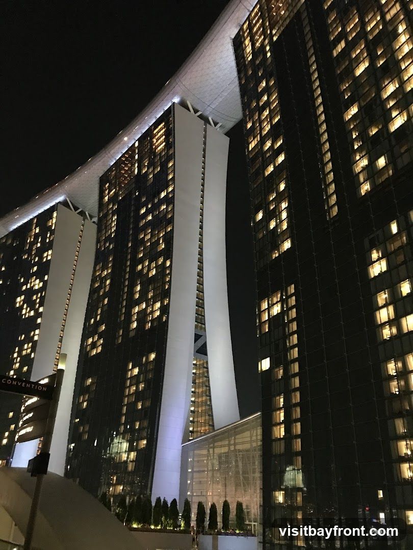 A nighttime view of the iconic Marina Bay Sands hotel in Singapore, showcasing its distinctive architecture with three towers connected by a sky-high platform. The buildings are illuminated with numerous lights from their windows, creating a striking contrast against the dark sky. A sign indicating "CONVENTION" is visible on the left side of the image.