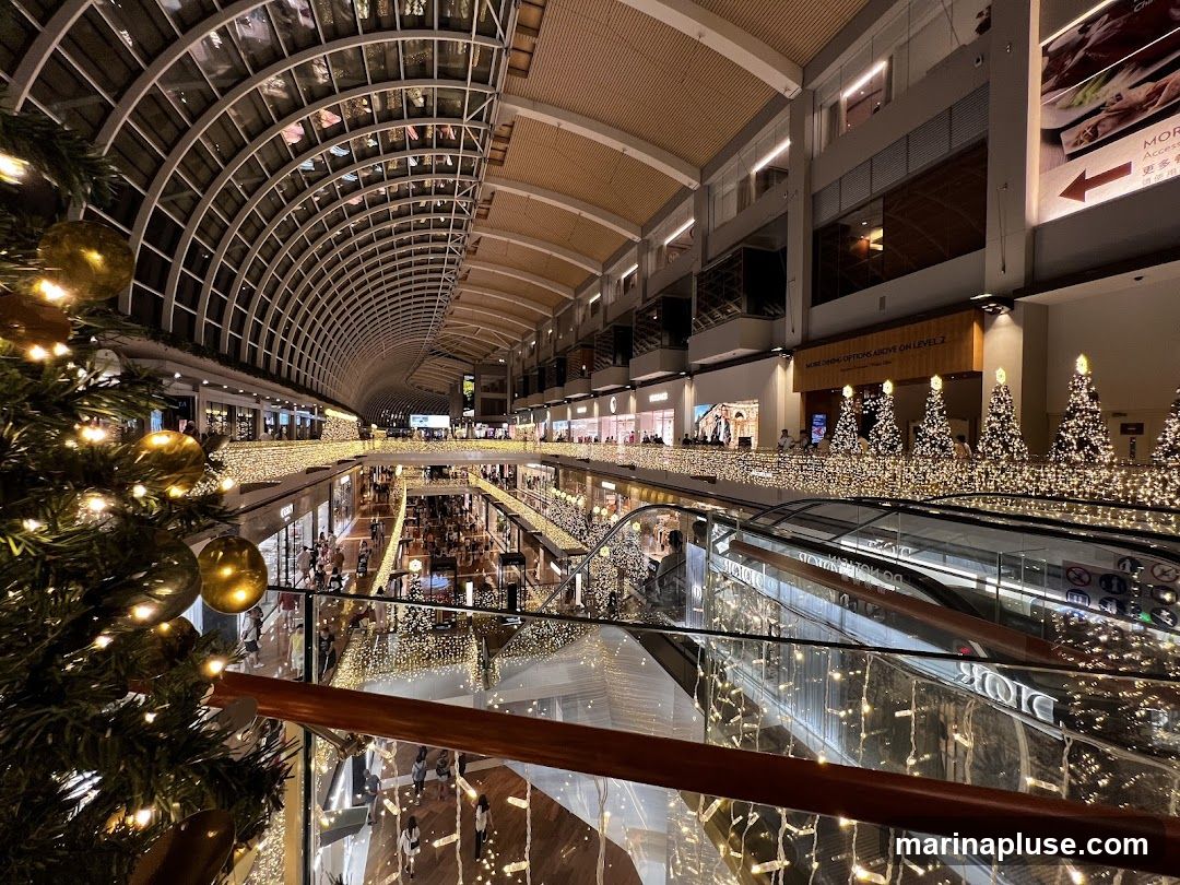 A wide-angle view of the interior of a multi-level shopping mall, adorned with extensive Christmas decorations. String lights are draped along railings and archways, and several decorated Christmas trees are visible. People are seen browsing shops and walking on escalators and walkways. The architecture features a large, curved glass ceiling.
