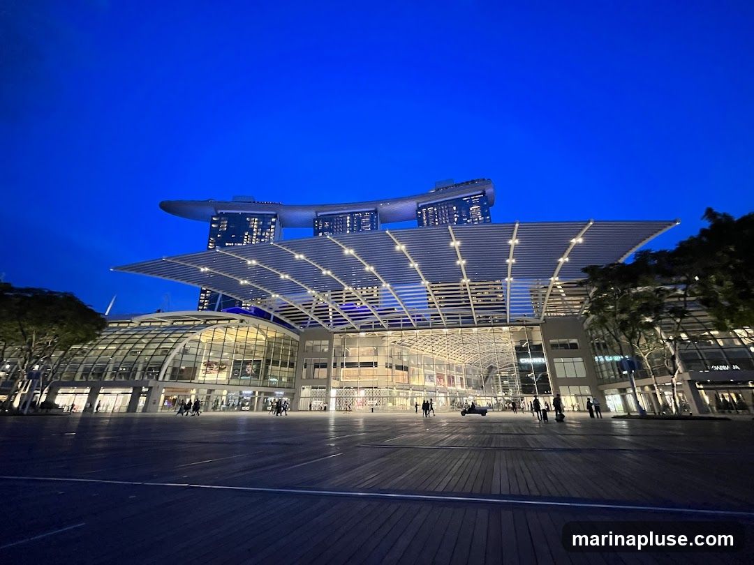 A wide shot of the Marina Bay Sands complex in Singapore at dusk. The iconic three-tower hotel with its sky park is visible in the background, illuminated against the deep blue sky. In the foreground, the convention center and shopping mall area features a modern architectural design with a large, cantilevered roof structure and extensive glass facades. People are seen walking around the plaza in front of the buildings. The wooden boardwalk in the foreground is lit, reflecting the ambient light.