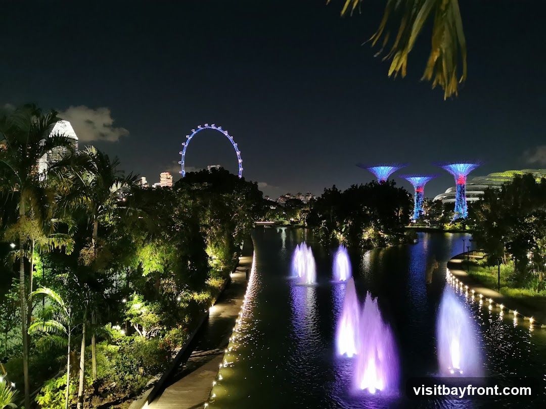 A nighttime scene at Gardens by the Bay, Singapore. In the foreground, illuminated fountains spray water in various colors, including purple and blue, across a dark body of water. The pathway along the water's edge is lined with lights. Lush greenery and palm trees are visible on the left, with some trees illuminated by spotlights. In the background, the iconic Supertrees are lit up in vibrant blue and red. Further in the distance, the Singapore Flyer Ferris wheel is visible, along with city skyline buildings. The sky is dark with a few clouds.