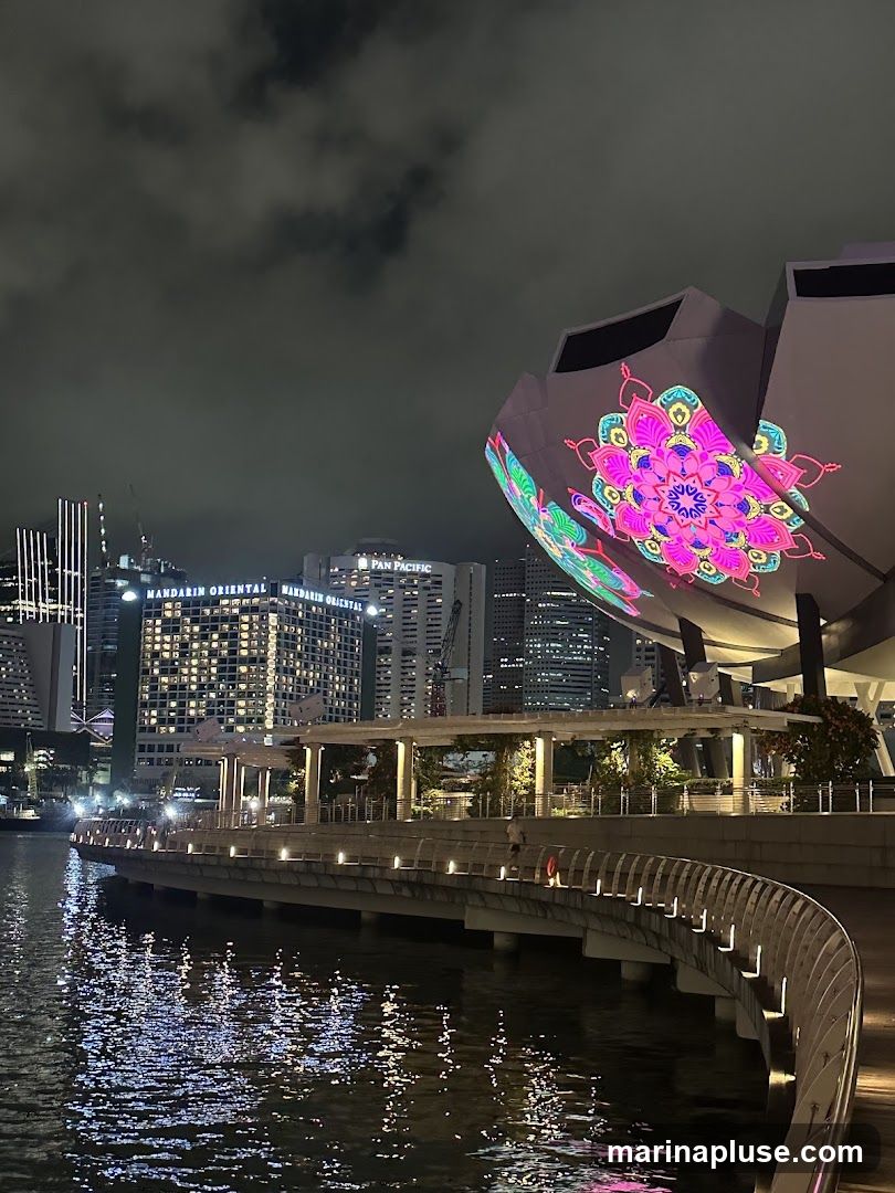 A nighttime view of a cityscape with a body of water in the foreground reflecting the lights. A curved walkway lines the water's edge, illuminated by small lights. In the background, several tall buildings are visible, including one with "MANDARIN ORIENTAL" and "PAN PACIFIC" signage. A distinctive building in the mid-ground is illuminated with a large, colorful mandala-like projection. The sky is dark and cloudy.