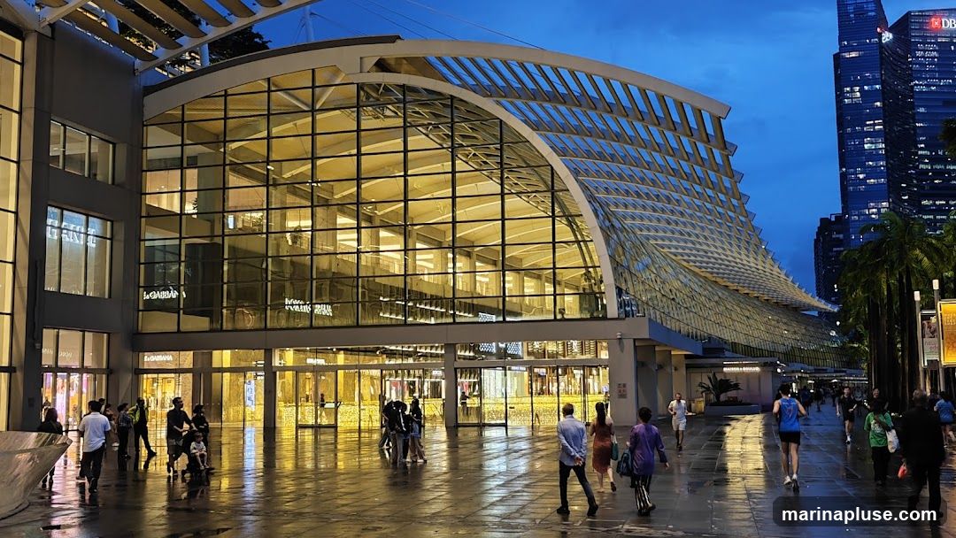 A wide shot of a large, modern building with a distinctive curved glass roof structure. The building's facade is predominantly glass, reflecting the warm interior lights and the twilight sky. People are walking on the wet pavement in front of the building, suggesting it is a public space or shopping area. In the background, tall city skyscrapers are visible. The overall scene captures an urban environment at dusk.