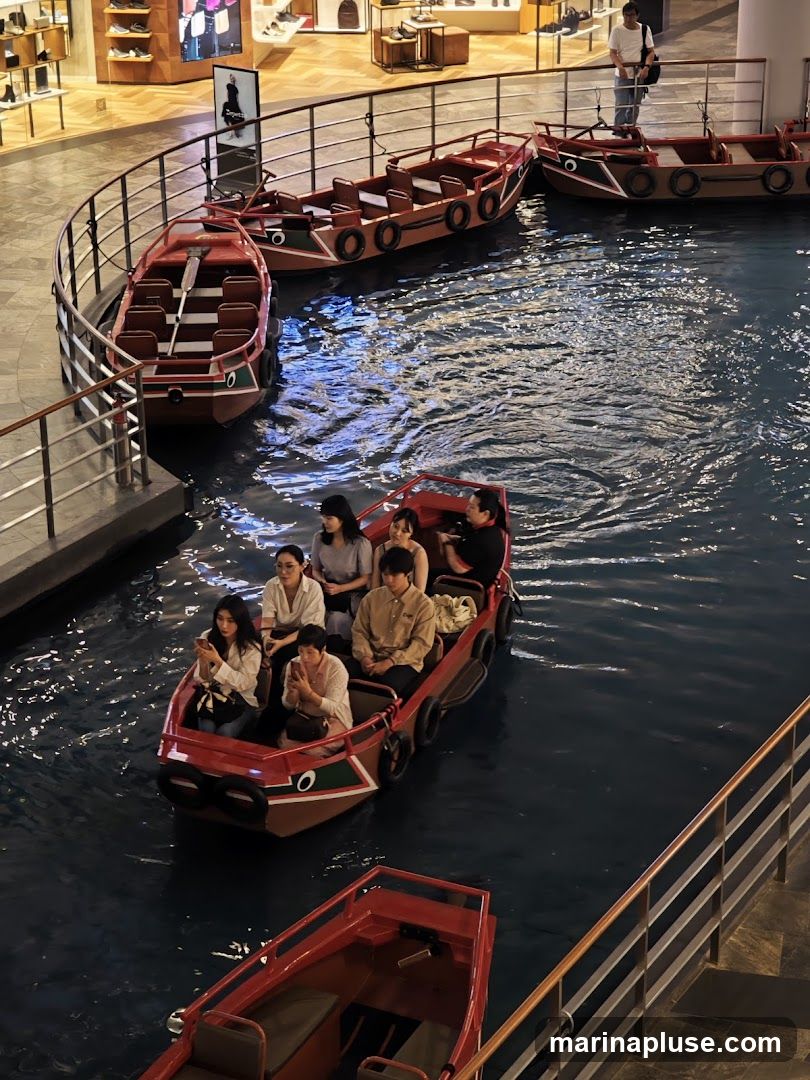 People are enjoying a gondola ride in an indoor setting, possibly a shopping mall. Several red and brown gondolas are on a dark blue water channel, with some occupied by passengers. In the background, there are shops with displays of merchandise.