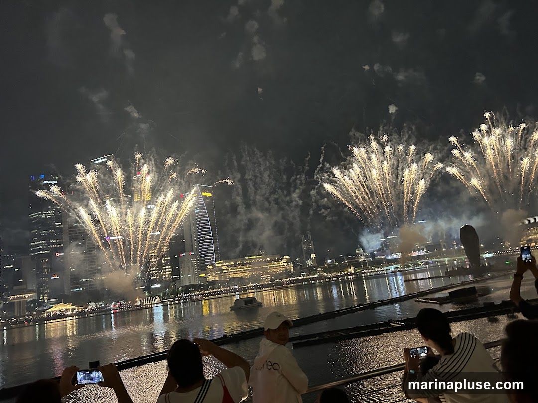 A vibrant fireworks display illuminates the night sky above a city skyline, with numerous buildings reflecting in the water below. People are gathered along a waterfront, many holding up phones to capture the spectacle. The fireworks are golden and white, fanning out in various patterns against the dark sky.