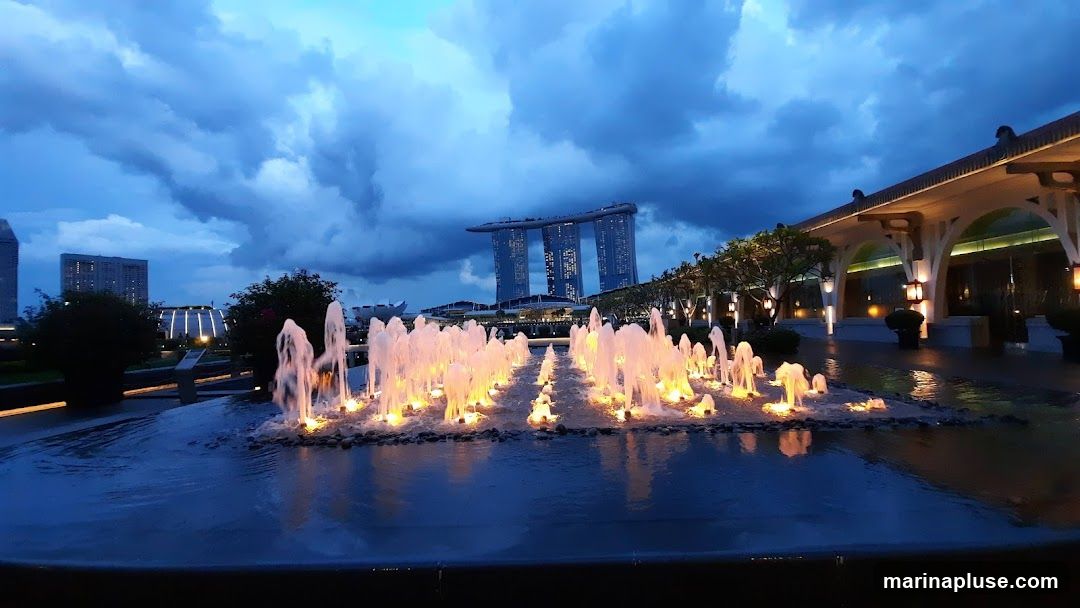 A wide shot of a water fountain display in front of a building with arched walkways and lanterns. In the background, the iconic Marina Bay Sands hotel is visible against a dramatic, cloudy sky at dusk. The water reflects the lights of the fountain and the surrounding architecture.