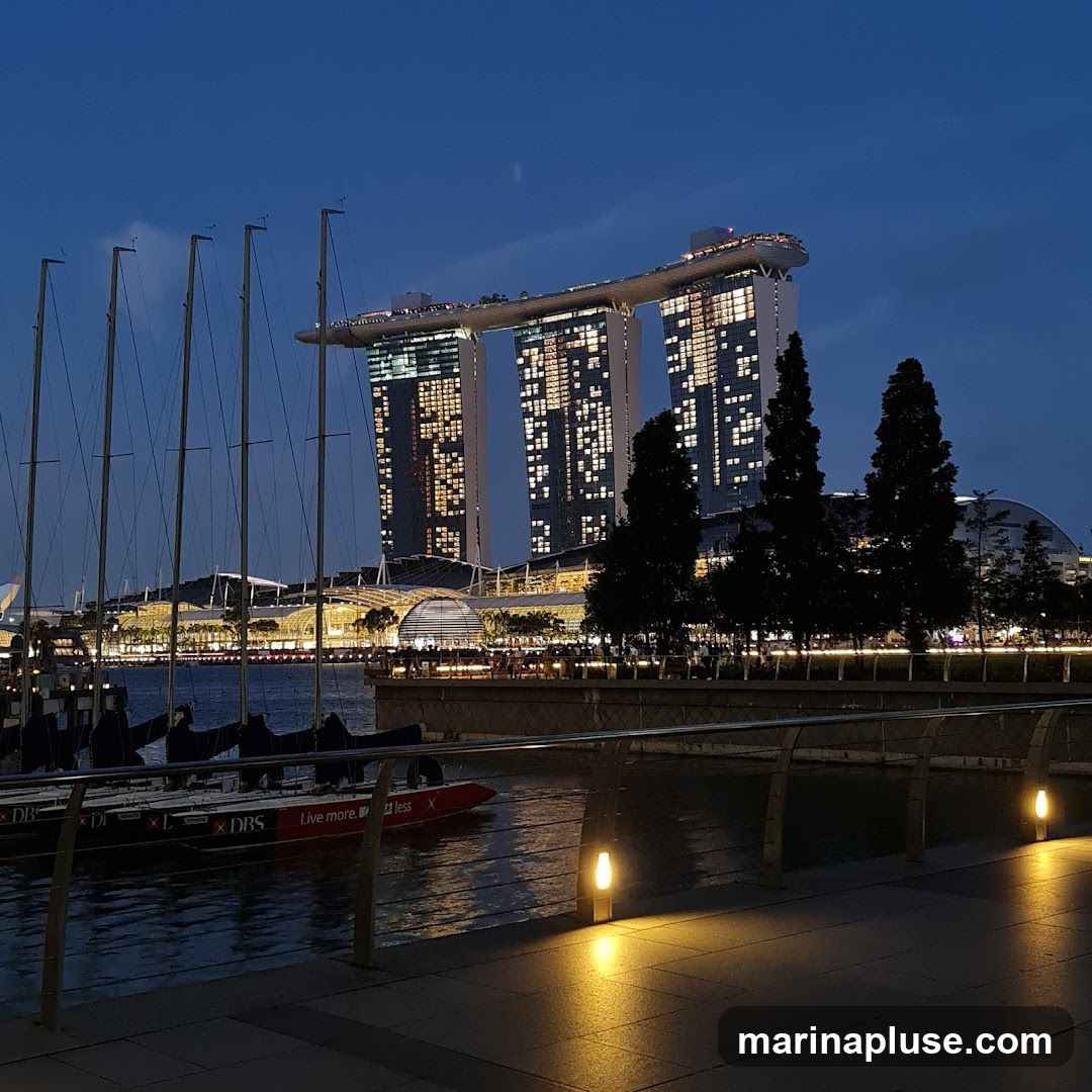 A wide shot of the Marina Bay Sands hotel in Singapore at dusk. The iconic building, with its three towers and sky park, is illuminated with many lights. In the foreground, several sailboats are docked in the water, their masts tall against the twilight sky. A walkway with modern railings and glowing lamps lines the waterfront, leading the eye towards the impressive cityscape.