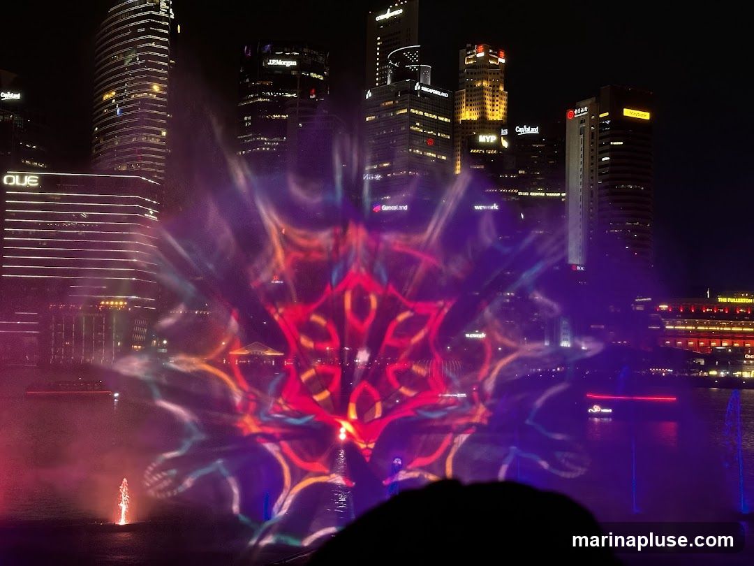 A vibrant light show is projected onto mist over a body of water at night. The projection features a large, colorful, mandala-like pattern in shades of red, orange, purple, and blue. In the background, a modern city skyline is illuminated with numerous skyscrapers. Some buildings have visible signage, including 'J.P. Morgan', 'MYP', 'CapitaLand', 'we work', and 'The Fullerton Hotel'. Water fountains are also visible in the foreground and mid-ground, illuminated by colored lights. The silhouette of a person's head is visible in the bottom foreground.