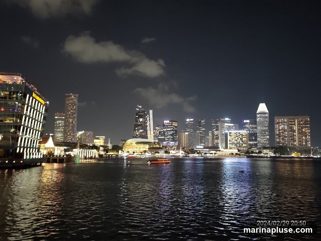 A nighttime view of a city skyline across a body of water. The buildings are illuminated with various lights, and there are clouds in the dark sky. Several boats are visible on the water. The date and time stamp '2024/02/29 20:50' is in the bottom right corner.
