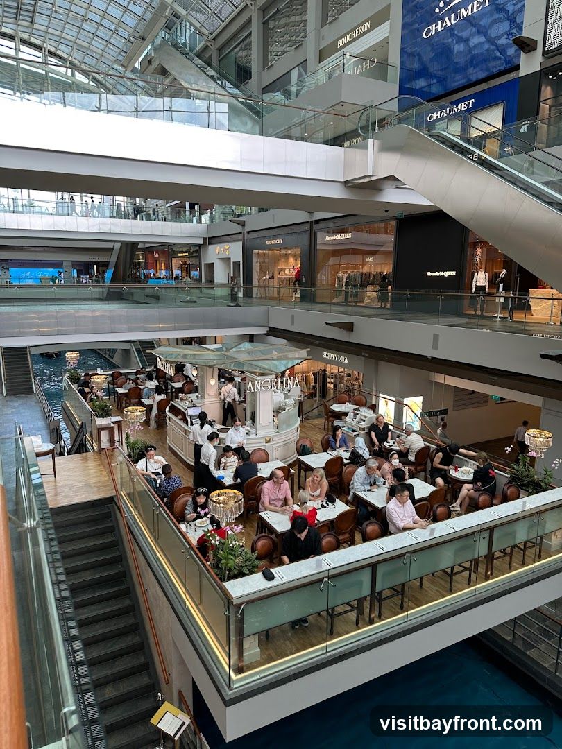 An interior view of a large, modern shopping mall with multiple levels. A canal or water feature is visible at the bottom. On one of the upper levels, a cafe named ANGELINA is bustling with customers seated at tables. Other shops, including luxury brands like GIVENCHY and ALEXANDER MCQUEEN, are visible on higher floors. Escalators and walkways connect the different levels.