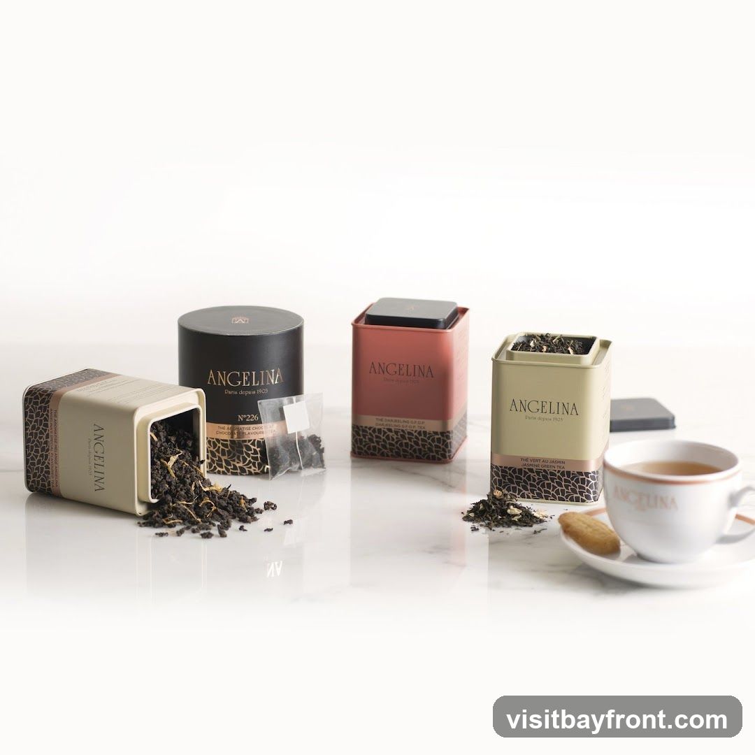 A still life arrangement featuring three decorative tins of Angelina tea, a cup of tea, and a biscuit on a white marble surface. One tin is lying on its side, spilling loose tea leaves. The tins are labeled with the Angelina brand and "Paris depuis 1903".