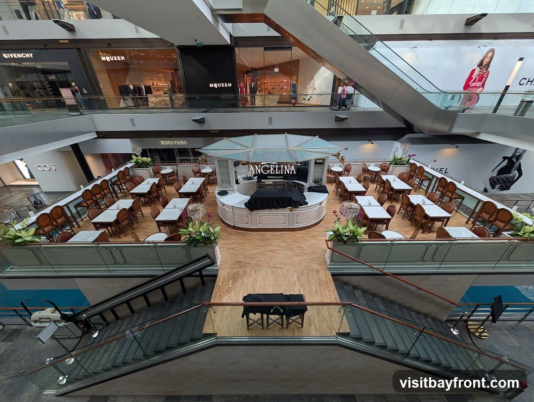 An overhead view of the seating area for the Angelina cafe, located within a multi-level shopping mall. The cafe features a central counter with the Angelina logo prominently displayed. Numerous tables and chairs are arranged on a raised platform, surrounded by railings. In the background, various retail stores like GIVENCHY, MCQUEEN, and COS are visible on different levels of the mall. Large advertisements for fashion brands are also present on the mall walls.