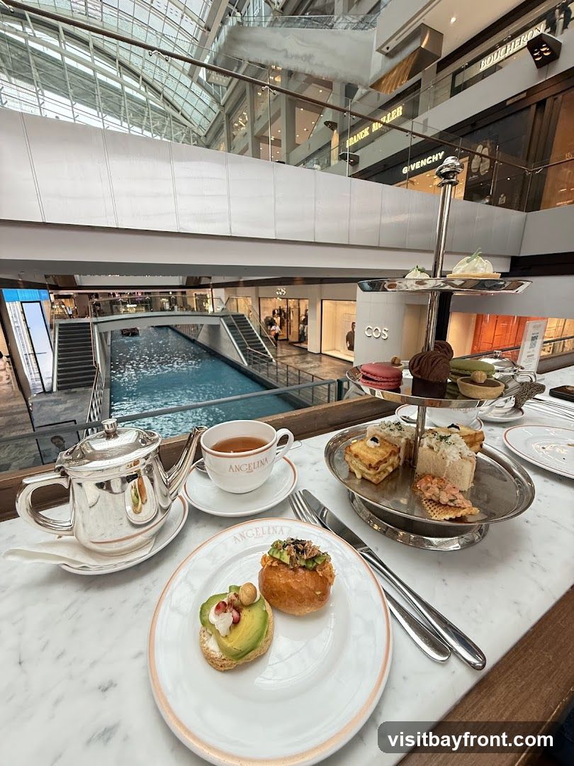 A table set for afternoon tea with a silver teapot, a cup of tea, and a tiered stand filled with pastries and sandwiches. The setting appears to be indoors within a large, modern shopping mall with a canal running through it. The Angelina logo is visible on the teacup and plate.