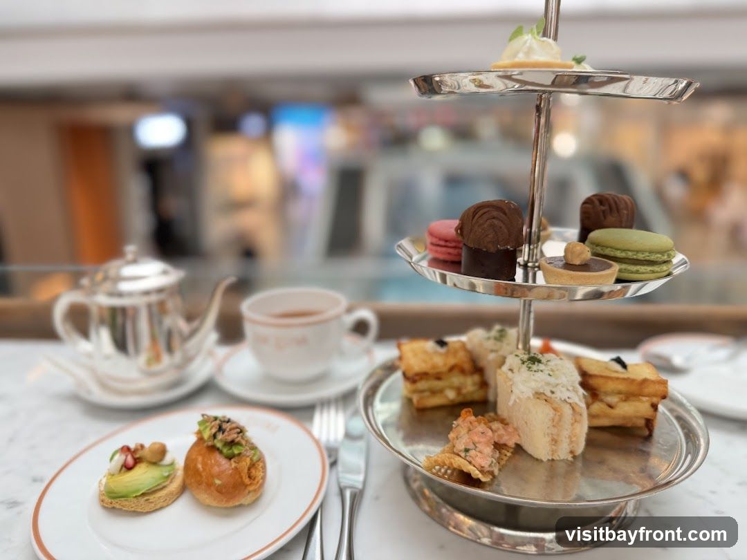 A tiered stand holds an assortment of delicate pastries and sandwiches, accompanied by a teapot, teacup, and a small plate with two appetizers. The background is blurred, showing what appears to be a shopping mall interior with escalators and illuminated displays.