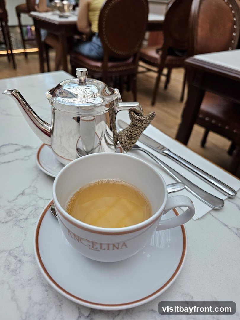 A close-up shot of a tea set on a marble-patterned table. A silver teapot and a white teacup with the word "ANGELINA" printed on it are in focus. The teacup is filled with a light-colored beverage, likely tea. Cutlery is visible to the right of the teacup. In the background, blurred figures and leather chairs suggest a dining or cafe setting.