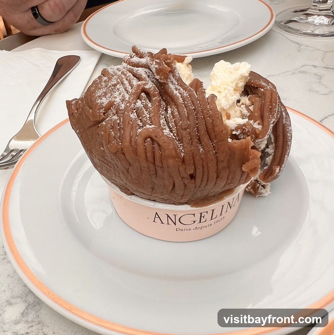 A close-up of a Mont Blanc dessert, a pastry made of chestnut puree, topped with whipped cream and dusted with powdered sugar. The dessert is served in a pink paper cup with the Angelina Paris logo. It sits on a white plate with a pink rim, next to a fork and a napkin.