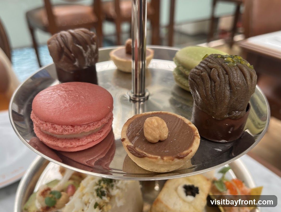 A close-up shot of a tiered serving tray filled with various small desserts. The top tier features a pink macaron, a chocolate tart topped with a hazelnut, a chocolate pastry resembling a mushroom, and a green macaron next to a dark chocolate pastry with green sprinkles. The lower tier, partially visible, shows savory appetizers including what appears to be a ceviche or tartare on a chip, and another appetizer with black caviar. The background is blurred, showing chairs and a table setting.