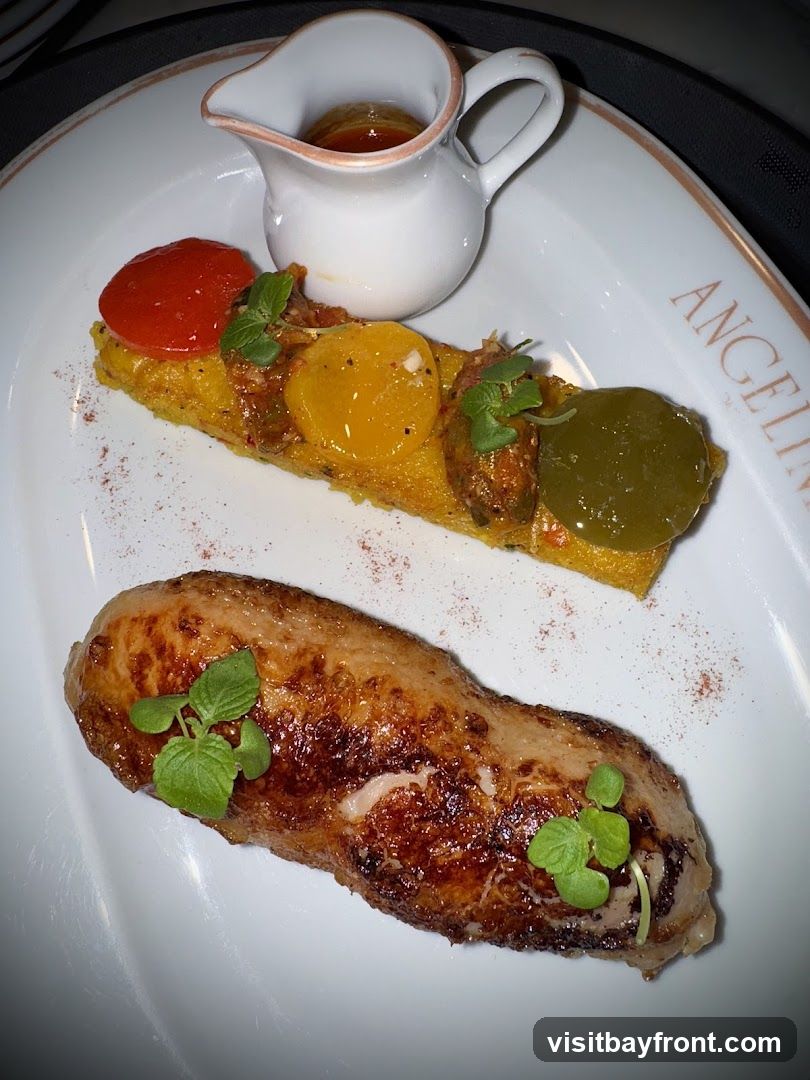 A white plate holds a gourmet dish featuring a piece of seared meat, possibly poultry, garnished with fresh green herbs. Alongside the meat is a rectangular portion of food topped with slices of red, yellow, and green vegetables, and a small white pitcher filled with a golden-brown sauce. The plate has a subtle embossed logo that reads "ANGELINA".